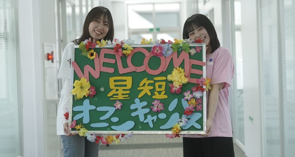WELCOME 星短 オープンキャンパスと書いたボードを見せる女子大学生2人
